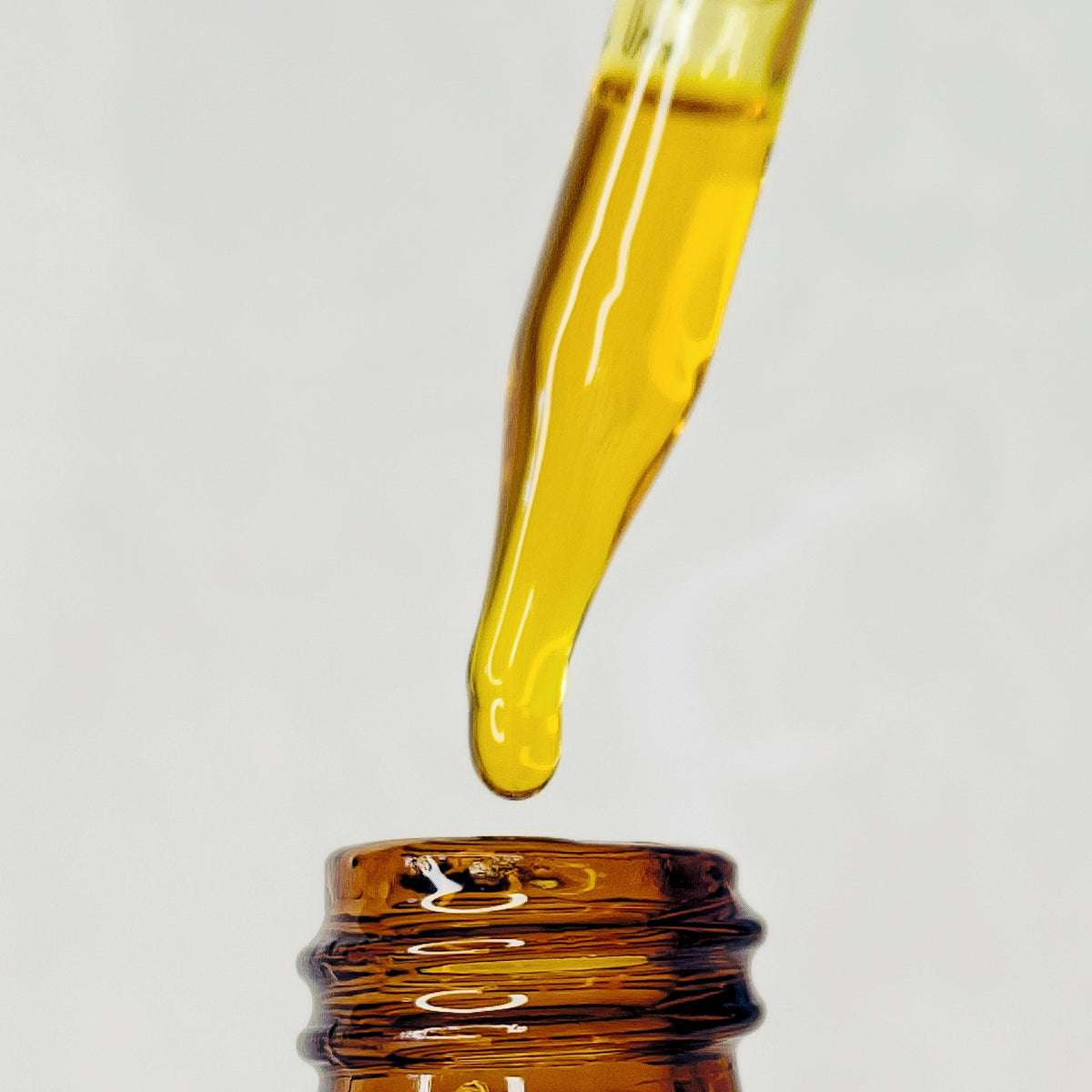Golden Harmony Serum facial oil forming a drop at the tip of a glass dropper above an amber bottle.