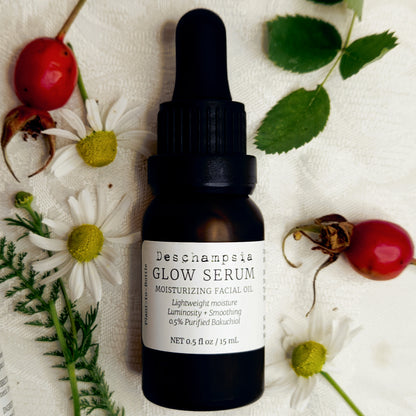 Deschampsia Glow Serum 15 mL dropper bottle surrounded by chamomile flowers, rosehips and rose leaves.