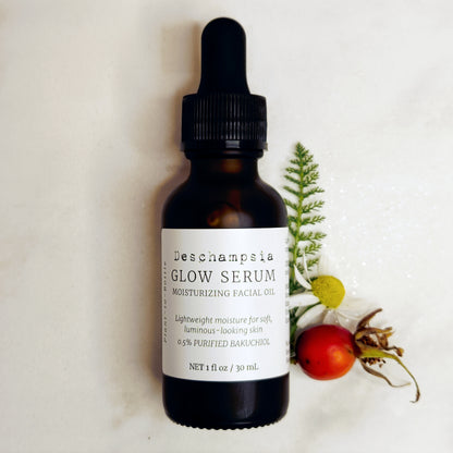 Deschampsia Glow Serum 30 mL amber dropper bottle beside a rosehip, chamomile flower and yarrow foliage on a cream background.