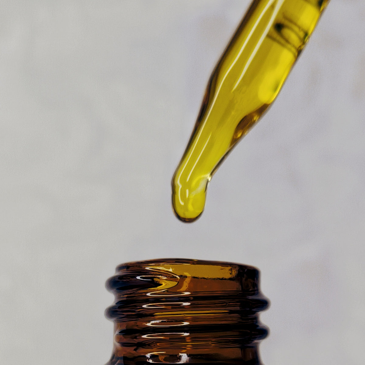 A drop of golden Glow Serum facial oil at the tip of a glass dropper above an amber bottle.