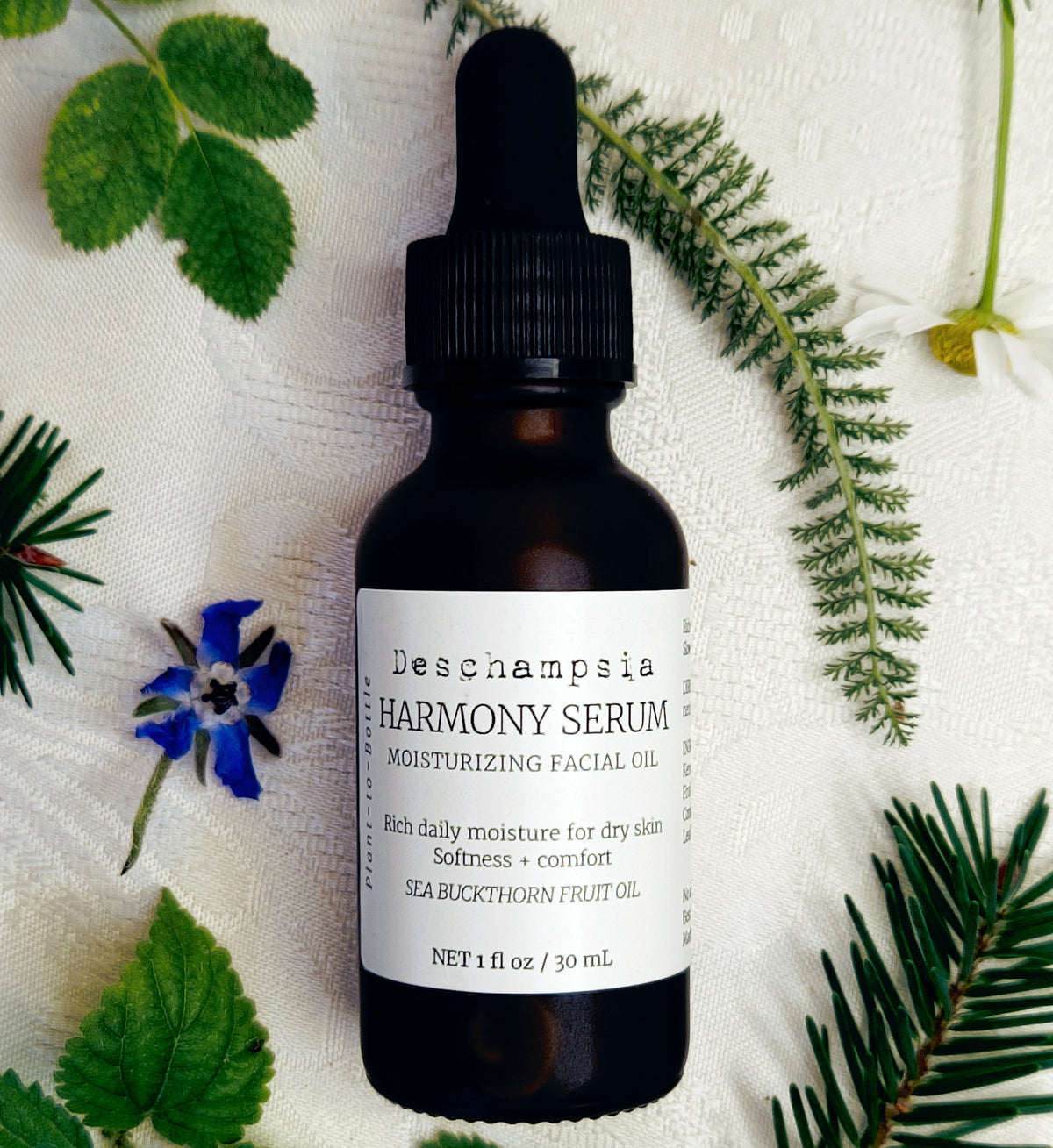 Deschampsia Harmony Serum 30 mL bottle with blue flowers, green leaves, fern and evergreen sprigs on cream fabric.