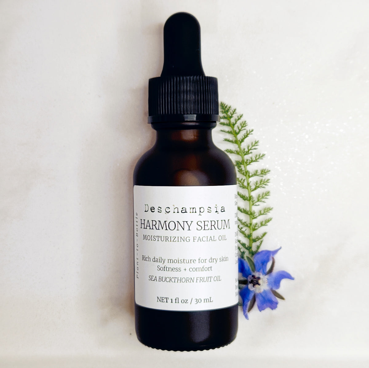 Deschampsia Harmony Serum 30 mL amber dropper bottle beside blue flowers and a fern frond on cream fabric.